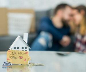 First-time buyer mortgage a toy house in fore with post it saying our first home, blurred couple in the background