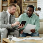 Two men sat on a sofa checking a calculator and paperwork